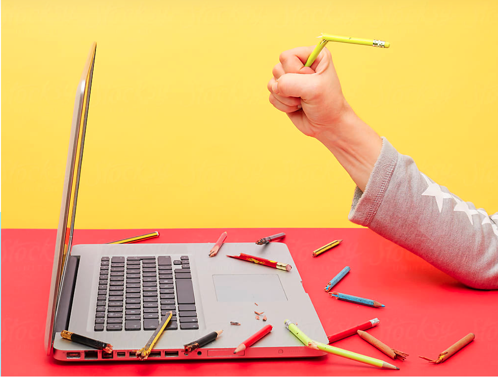 A frustrated hand grasps a broken pencil in front of a laptop
